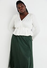 Blusa bianca plissettata con scollo a V profondo e maniche lunghe, abbinata a una gonna plissettata fluente di un verde scuro, caratterizzata da una texture diagonale.
