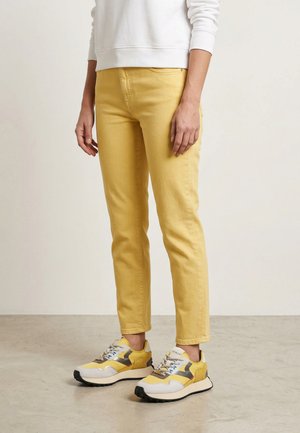 Osoba v žlutých úzkých kalhotách, bílém svetru a žluto-bílých teniskách stojící na světle šedé podlaze.