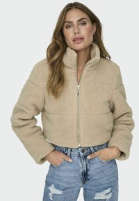 Giacca in pile beige tagliata con colletto alto, zip frontale e superficie testurizzata; abbinata a jeans blu strappati.