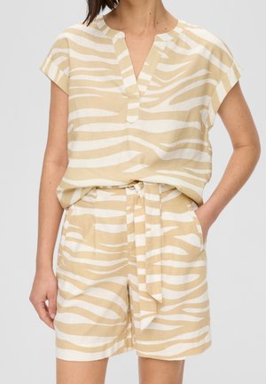 Vrouw draagt een beige-witte blouse met zebraprint en korte mouwen en bijpassende korte shorts met hoge taille en stoffen riem, één hand in de zak.