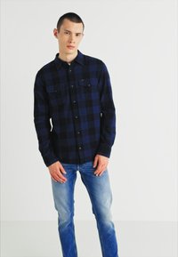 Cămașă de flanel în carouri navy și negre, cu închidere cu nasturi și două buzunare pe piept, asociată cu blugi din denim ușor albastri.