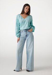 En lyseblå, struktureret bluse med blomsterprint og lange ærmer, parret med høj talje, vide lys jeans og sorte hæle.