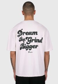 White cotton t-shirt with short sleeves, featuring bold black text on the back: "Dream Big, Grind Bigger" with "Dropsize" underneath.
