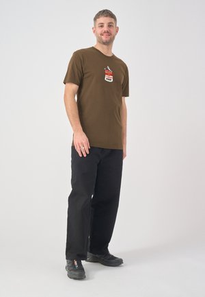 Mann steht lächelnd da, trägt ein braunes T-Shirt mit Suppe-Dosen-Grafik, schwarze Hose und schwarze Turnschuhe vor schlichtem Hintergrund.
