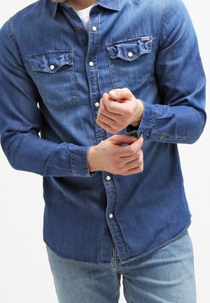 Blue denim shirt with long sleeves, two chest pockets, button-down collar, and a slim fit design. Fabric appears soft with a clean finish.