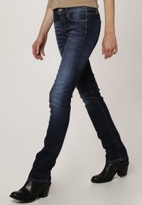 Jeans ajustados de mezclilla en azul oscuro, con un diseño clásico de cinco bolsillos y un ligero desgastado. Combinados con botines negros de tacón.