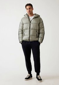 Lichtgroene gewatteerde jas met capuchon, voorzien van een ritssluiting en zijzakken. Gecombineerd met zwarte trainingsbroek en zwarte sneakers.