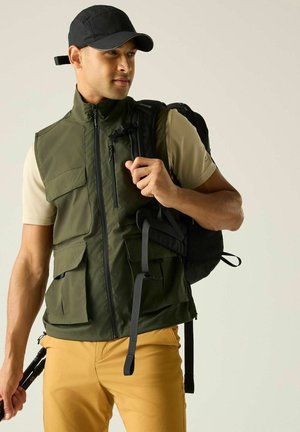 Hombre con gorra negra, chaleco verde, sujetando la correa negra de una mochila y bastones de senderismo, vistiendo camisa beige y pantalones mostaza sobre fondo liso.