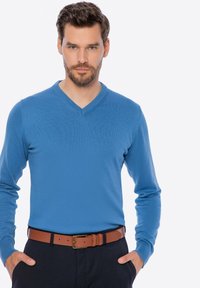 Niebieski sweter w serek wykonany z gładkiego dzianinowego materiału, z długimi rękawami. Nosi się go z brązowym skórzanym paskiem na ciemnych spodniach.