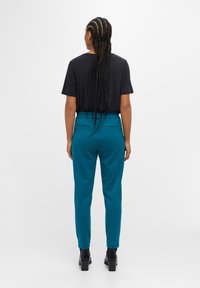 Femme aux longs cheveux tressés, vue de dos, portant un haut noir à manches courtes, un pantalon bleu sarcelle et des bottines noires à talons.