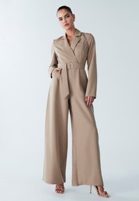 Bēša piemērota kombinezona ar garām piedurknēm, jostu viduklī un platām biksēm. Gluda auduma ar smalku spīdumu un izgriezuma atloku.