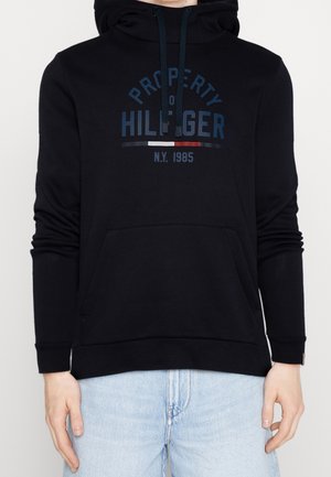 Černá mikina s kapucí, přední kapsou, stahovací šňůrkou a nápisem s logem „Property of Hilfiger N.Y. 1985“ uprostřed na hrudi, nošená s světle modrými džínami.