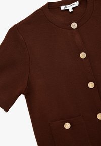 Cárdigan de punto marrón con mangas cortas, que presenta dos bolsillos frontales y cierres de botón dorados. Tejido texturizado con bordes acanalados.