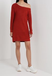 Rotes, gestricktes Kleid mit asymmetrischem One-Shoulder-Design, langen Ärmeln und lockerer Passform, kombiniert mit weißen Sneakers.