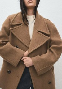 Cappotto marrone in misto lana con design a doppiopetto, ampi revers e grandi bottoni. Presenta una vestibilità ampia con maniche oversize.