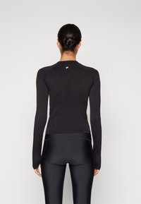 Svart långärmad träningströja med en figursydd design, texturerad bakpanel och en elegant silhuett, i kombination med svarta leggings.