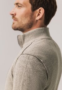 Grauer Sweatshirt mit Reißverschluss und hohem Kragen, ausgestattet mit einer weichen Textur, sichtbaren Nähten und einem figurbetonten Design, seitlich gesehen.