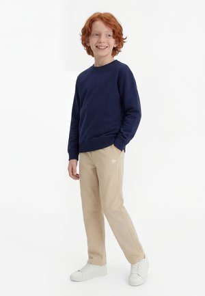 Raudonplaukis berniukas šypsosi, dėvi tamsiai mėlyną džemperį, smėlio spalvos kelnes su maža logotipu ir baltus sportinius batelius, stovi su viena ranka kišenėje.