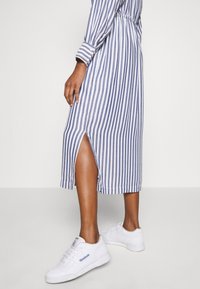 Gestreiftes Midikleid in Blau und Weiß mit einem seitlichen Schlitz und tailliertem Bund. Kombiniert mit weißen Sneakers, die einen lässigen Stil präsentieren.