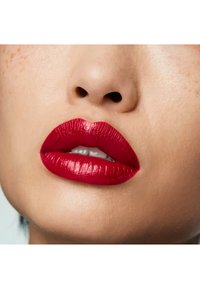 Rouge à lèvres rouge vif sur des lèvres plus pulpeuses, avec une finition brillante et une texture lisse. Le teint est uniforme avec des taches de rousseur discrètes autour du nez.