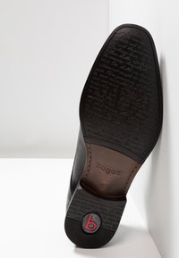 Chaussure en cuir noir avec une finition lisse et une semelle en caoutchouc. La semelle présente un texte en relief et un accent de logo rouge avec la lettre "b".
