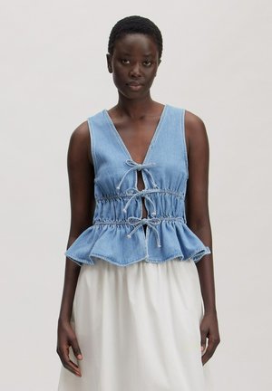 Woman with short hair wearing sleeveless blue denim peplum top tied with three bows and white skirt against plain background.