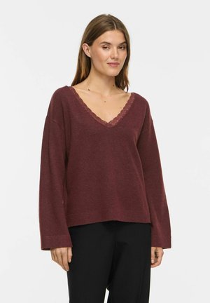 Femme portant un pull bordeaux à manches longues avec col en V et bordure en dentelle, ainsi qu'un pantalon noir, debout devant un fond blanc uni.