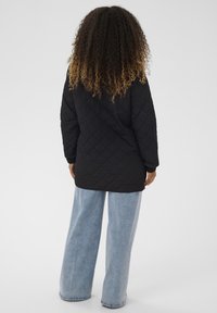 Nainen, jolla on kiharat ombre-hiukset, musta tikattu takki ja leveälahkeiset vaaleansiniset farkut, seisoo selin valkoista taustaa vasten.