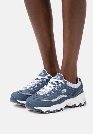 Personne portant des baskets Skechers D'Lites bleues et blanches avec des lacets blancs, debout sur un fond blanc.