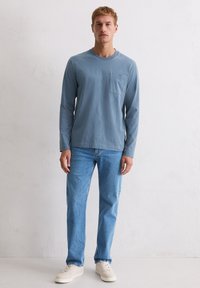 Jasnoniebieska koszula z długim rękawem i kieszenią z przodu, w połączeniu z jasnoniebieskimi dżinsami i beżowymi trampkami. Gładka bawełniana tkanina, luźny krój.