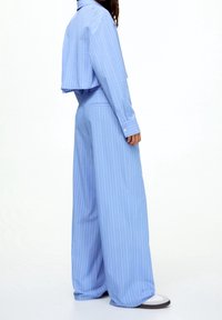 Osoba ubrana w jasnoniebieskie spodnie w paski w szerokim kroju oraz dopasowaną kurtkę, stojąca na tle jednolitego białego tła.