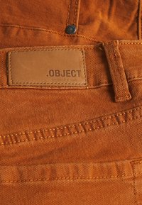 Primer plano de unos jeans de mezclilla color óxido mostrando la etiqueta de cuero de la marca que dice ".OBJECT artículo de coleccionista" y el detallado de las costuras.