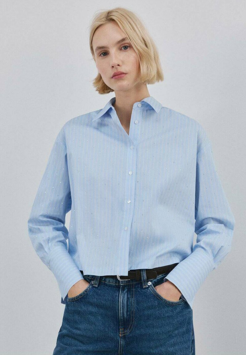 Chemise bleu clair à boutons avec des rayures verticales blanches et de petits points décoratifs. Manches longues et col classique. Portée avec un jean taille haute.