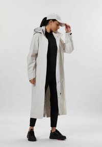 Ljusbeige lång kappa med huva, har tryckknappslås och manschettdetaljer, bärs över en svart topp och åtsittande byxor, kompletterad med en vit keps och svarta sneakers.