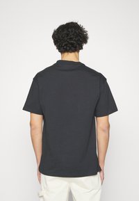 Personne aux cheveux bouclés et foncés portant un t-shirt noir uni à manches courtes et un pantalon clair, de dos contre un fond blanc.