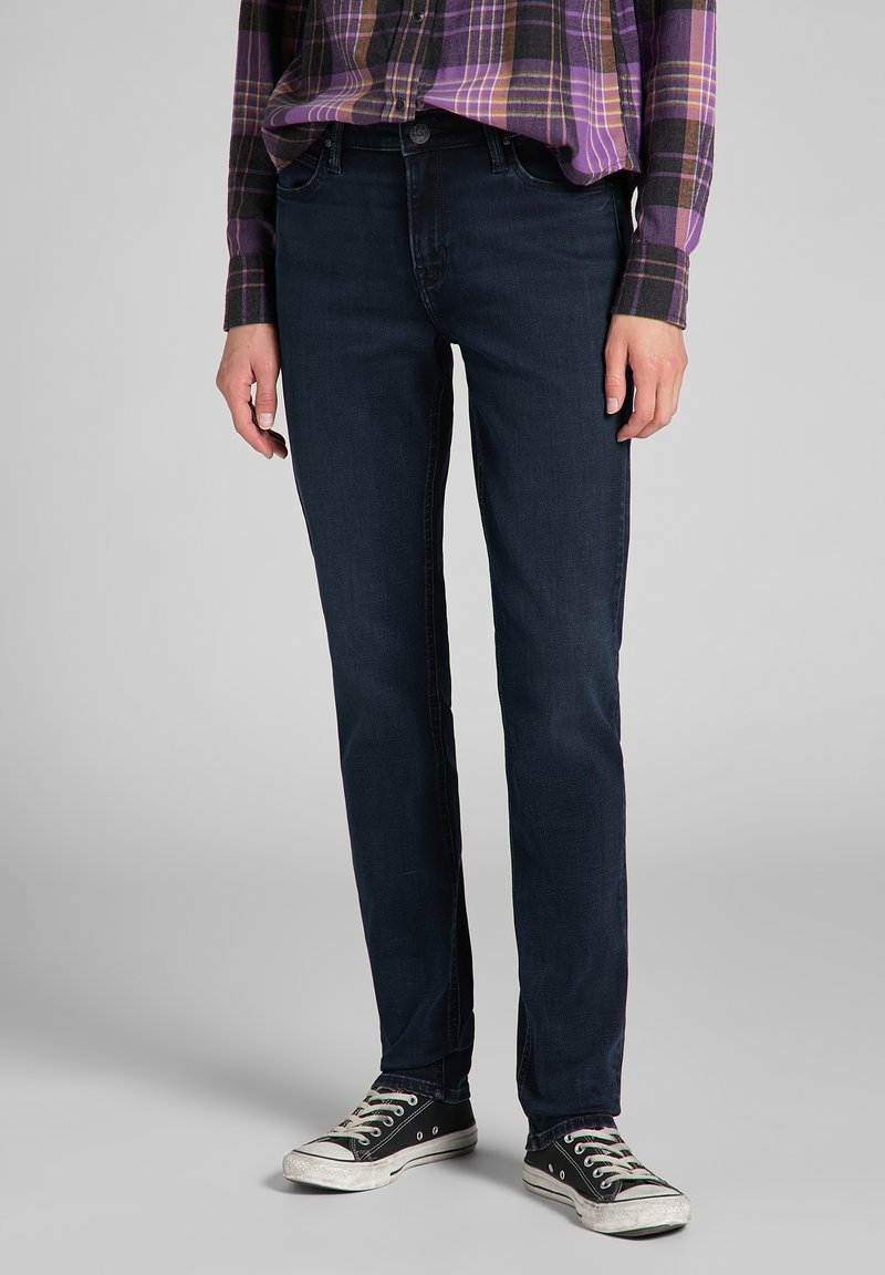 Jeans en denim bleu marine avec une coupe slim, présentant un design classique à cinq poches et des détails de couture subtils. Assortis à des baskets noires.