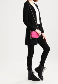 Černý kardigan, bílý top, černé legíny, černé šněrovací boty a malá růžová kabelka přes rameno. Zaměření na textury a kontrastní barvy.