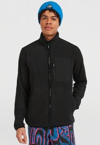 Giacca nera con zip e colletto alto, dotata di tasca sul petto, caratterizzata da una texture liscia. Indossata con pantaloni a motivi vivaci e un berretto blu.