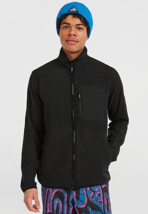 Zwarte zip-up jas met een hoge kraag en een borstzak, met een gladde textuur. Draagt zich met levendige, patroonrijke broek en een blauwe beanie.