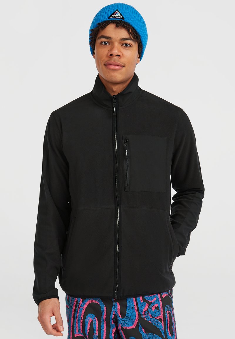 Giacca nera con zip e colletto alto, dotata di tasca sul petto, caratterizzata da una texture liscia. Indossata con pantaloni a motivi vivaci e un berretto blu.
