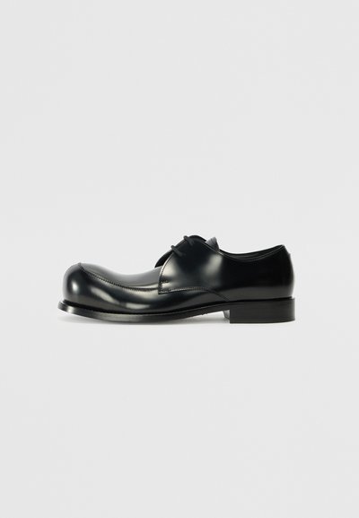Chaussure en cuir noir avec une finition brillante, bout arrondi, couture minimale et talon plat. Présente un système de laçage et une texture lisse.