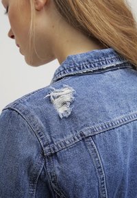 Žena s dlouhými blond vlasy, oblečená v modré džínové bundě s roztržením na horní části zad poblíž límce.