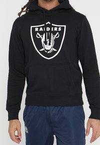 Felpa nera con cappuccio e tasca frontale, caratterizzata da un grande logo bianco dei Raiders e da un testo sovrastante. Il materiale sembra morbido con un design classico e aderente.
