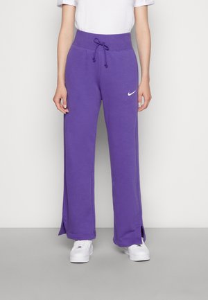 Personne portant un pantalon de survêtement violet avec cordon, fentes latérales et logo Nike blanc, associé à des baskets blanches et un t-shirt blanc à manches courtes.