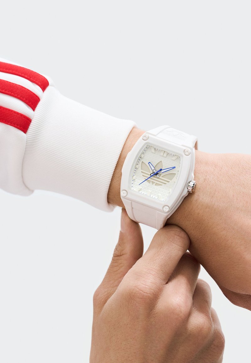 Orologio in silicone bianco con quadrante quadrato, dial testurizzato, lancette blu e logo Adidas. Indossato al polso, con le dita che regolano il cinturino.