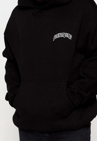 Sweat à capuche noir en tissu doux, avec une poche avant et un logo texte blanc. Comprend une capuche avec cordon de serrage et des poignets côtelés.