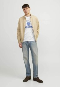 Beige zip-up takki valkoisen t-paidan päällä, jossa on sininen graafinen kuvio, yhdistettynä vaaleansiniset suorat farkut ja tummanruskeat kengät.