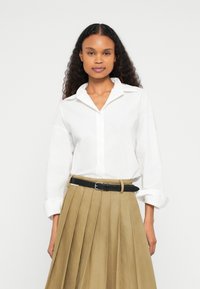 Camisa branca de botão com gola, mangas compridas e punhos dobrados. Combina com uma saia plissada bege e um cinto preto.