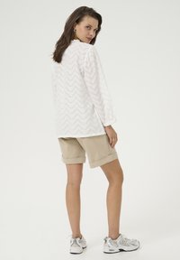 Chemise à manches longues à motifs de chevrons blancs, shorts en velours côtelé beige et baskets grises. La chemise présente un col volanté ; les shorts sont retroussés au niveau de l'ourlet.