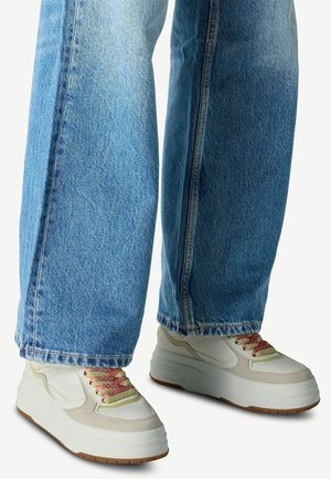 Nogi w jasnoniebieskich szerokich jeansach i białych sneakersach na platformie z różowymi i żółtymi sznurowadłami na jednolitym tle.